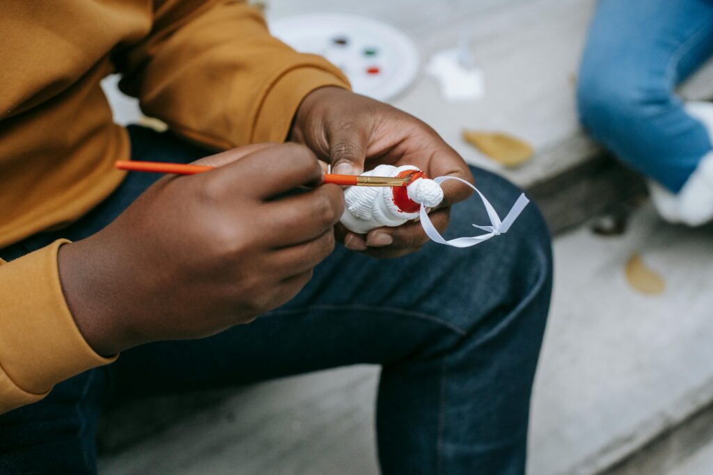 Unrecognizable African American male painting with paintbrush while decorating Christmas tree toy and preparing for holiday celebration on terrace with blurred background